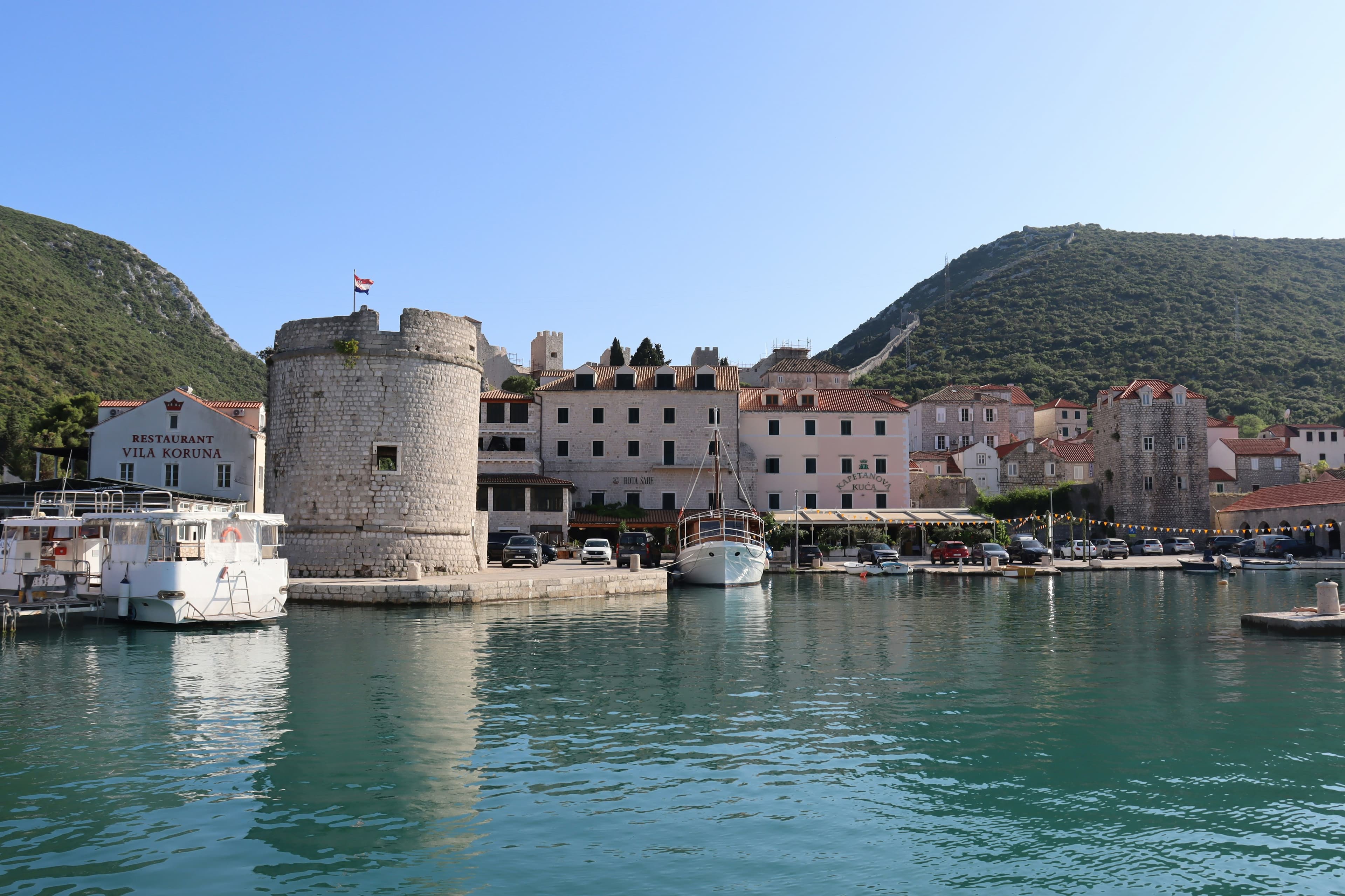 Mali Ston village waterfront with the round watchtower, the Bota Šare and Kapetanova Kuća restaurants and harbour-front buildings reflected in the still water.